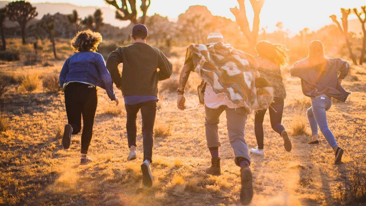 Four young adults running away from the camera through the desert, towards cacti and large rocks