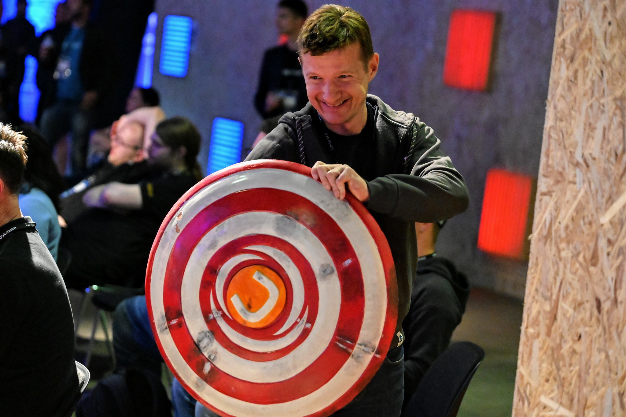 A man holding a Captain America-style shield with an orange Umbraco logo in the centre.